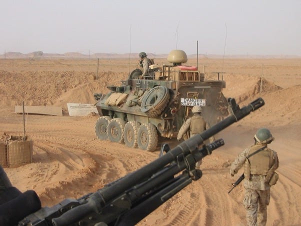 This color photo is of a Marine patrol departing the compound in remote area of al Anbar, Iraq in 2006.  The background terrain is dry, dusty predominantly flat with low mountains in the horizon. In the center is the back of an 8-wheeled armored logistics vehicle heading out of the compound. The vehicle has external storage, including spare tires and equipment, and a sign on the back that reads "DANGER STAY BACK."  Two soldiers on foot in full desert camouflaged combat gear are walking behind the armored vehicle.  In the foreground, a mounted machine gun is visible, suggesting the perspective is from someone inside or near a military vehicle. In the back of the image near the compound gate is a warning sign in the background reads "STOP PROCEED NO FURTHER.”