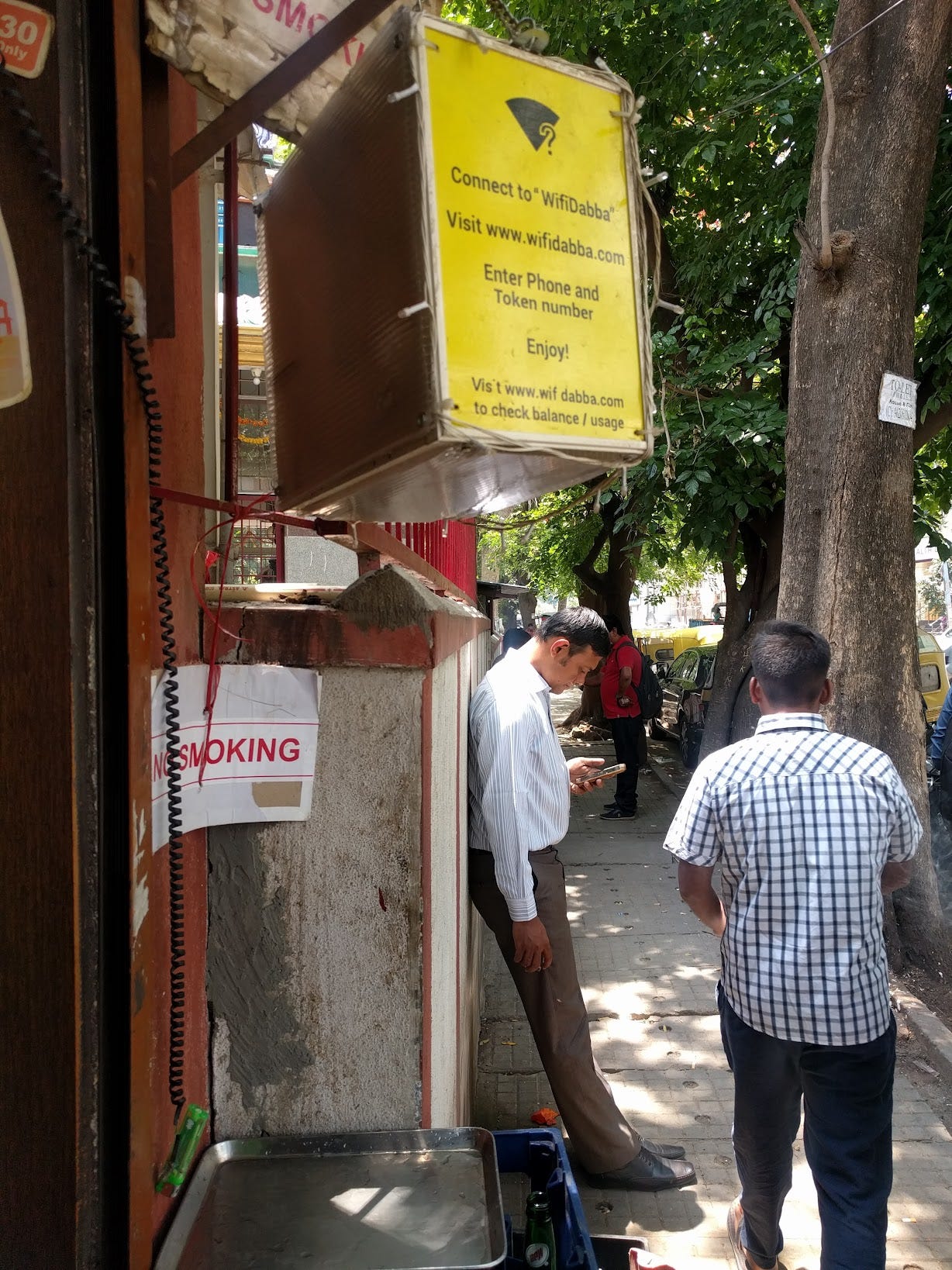 A busy street in India, with a man leaning against a wall checking his phone. A yellow sign above a kiosk advertises a wifi connection from a company called WifiDabba