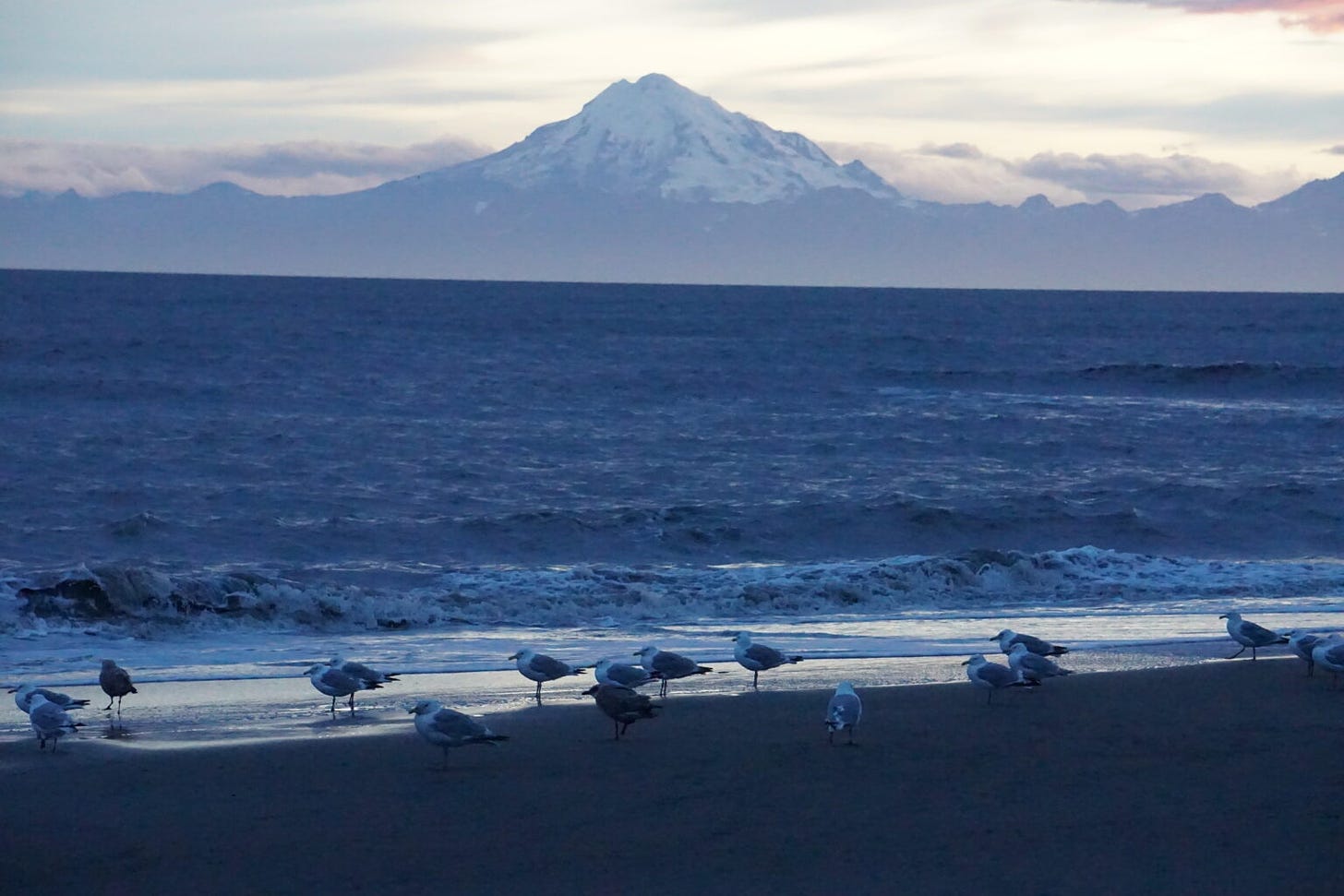 Waves lap on the Cook Inlet beach at Kenai on Aug. 14, 2018, with Redoubt Volcano looming over the opposite shore. The Cook Inlet region is teeming with renewable energy sources, including tidal and geothermal energy, experts say. (Photo by Yereth Rosen/Alaska Beacon)