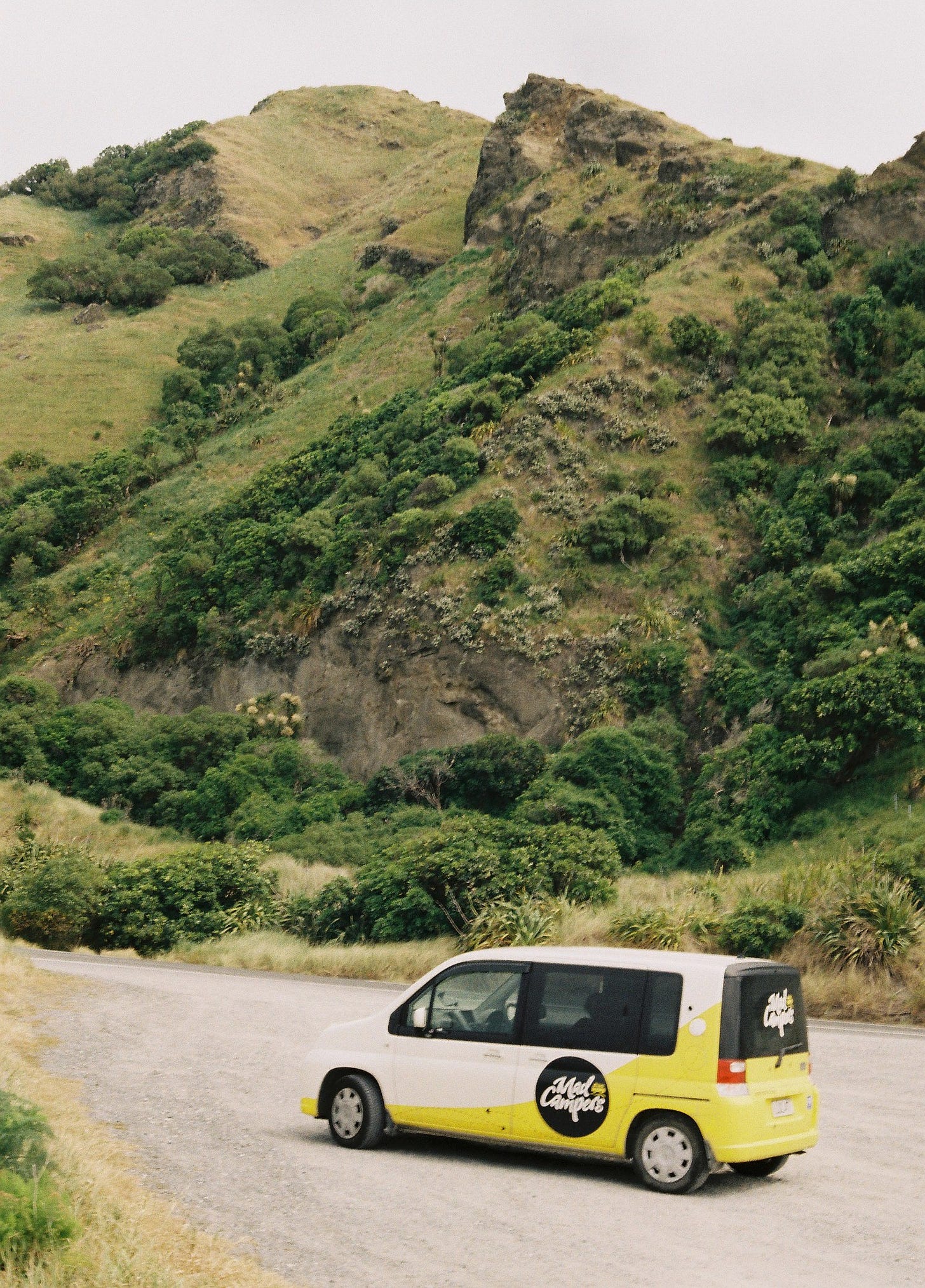 A small yellow and white camper van is parked beside a road and behind it, you can see a green hill