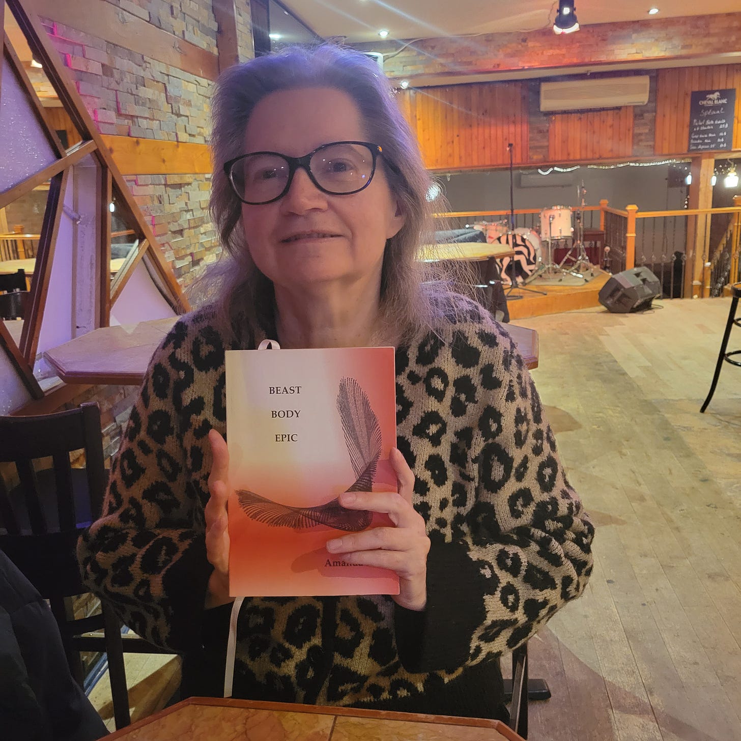 Amanda, wearing a leopard print sweater, sits in a bar at a table and holds a copy of Beast Body Epic, a book with red and black cover and a visual poem.