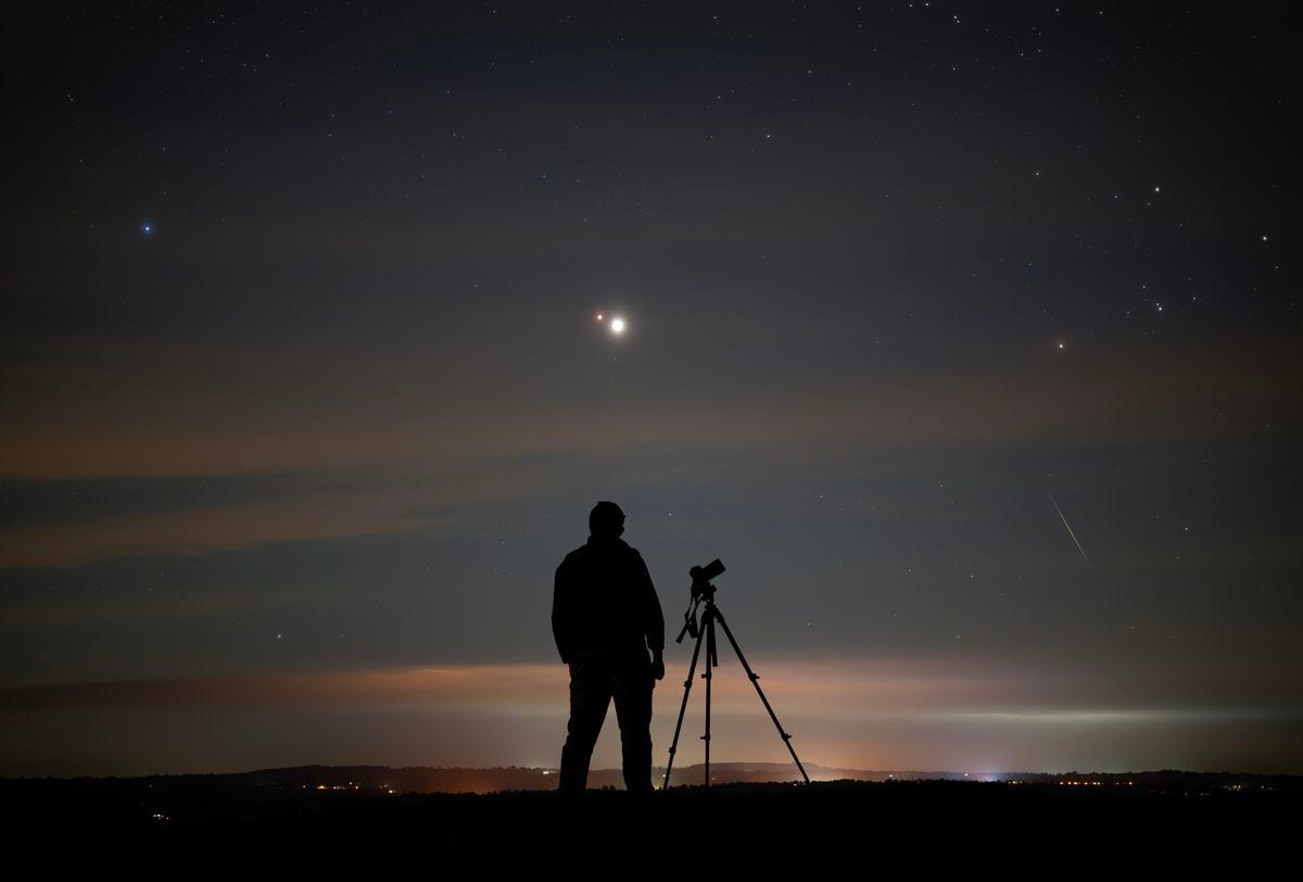 A dark figure is sillhouetted against a dark sky, with two bright lights--Jupiter and Mars--shining in the sky and a small meteor entering the sky on the right hand side. 