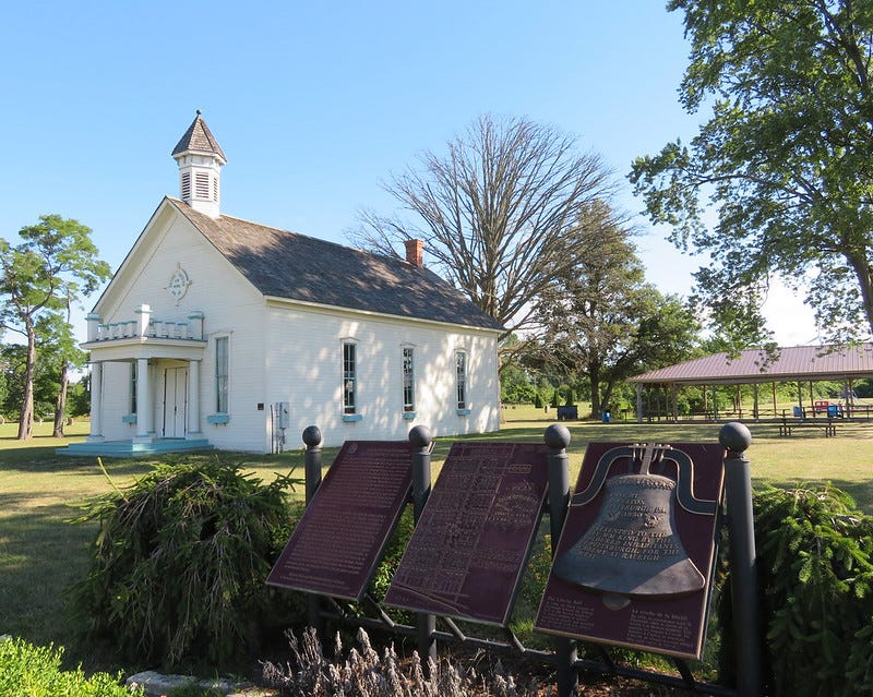 A white wooden building with a slate roof and a small bell tower. There is a porch of stone pillars at the front door, and three tall windows along the side. Details of the building are painted light blue. In the foreground is an information board that includes the image of a bell.