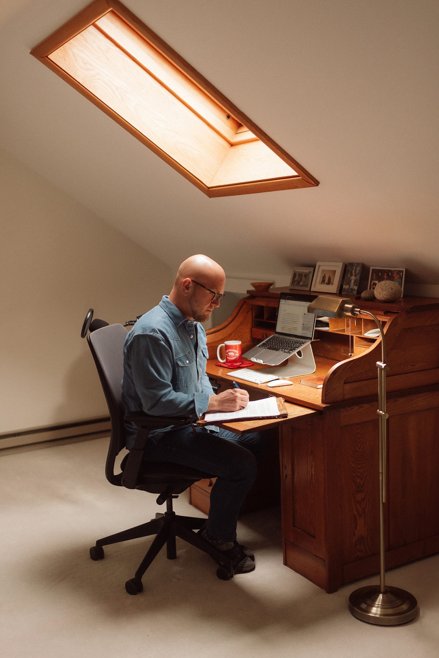 A writing coach sits at a wooden desk in a room with a skylight, working on a laptop with a notebook and pen. Framed photos are placed on the desk.