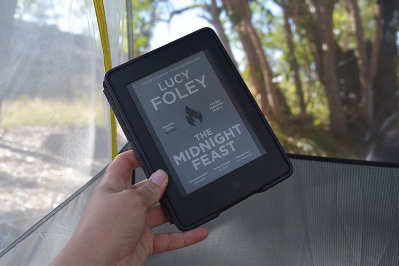 Hand holding the book ‘The Midnight Feast by Lucy Foley’ on Kindle in a tent.