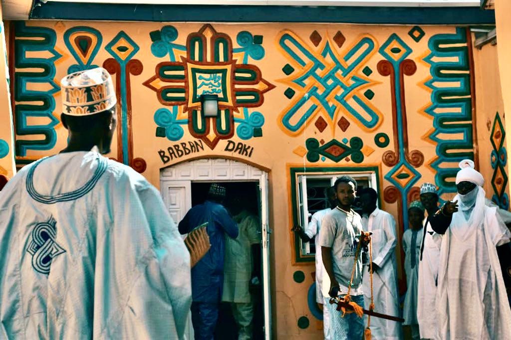 A building painted with beautiful blue, red and yellow geometric patterns, and a number of men in traditional Nigerian dress standing in front of it