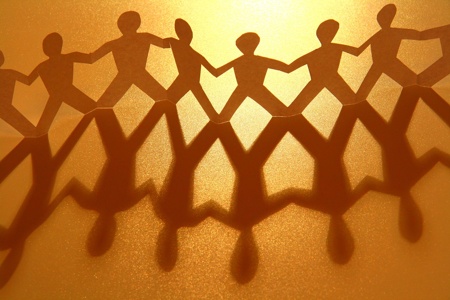 A line of connected paper cut-out people with a golden light shining from the background casting a long shadow