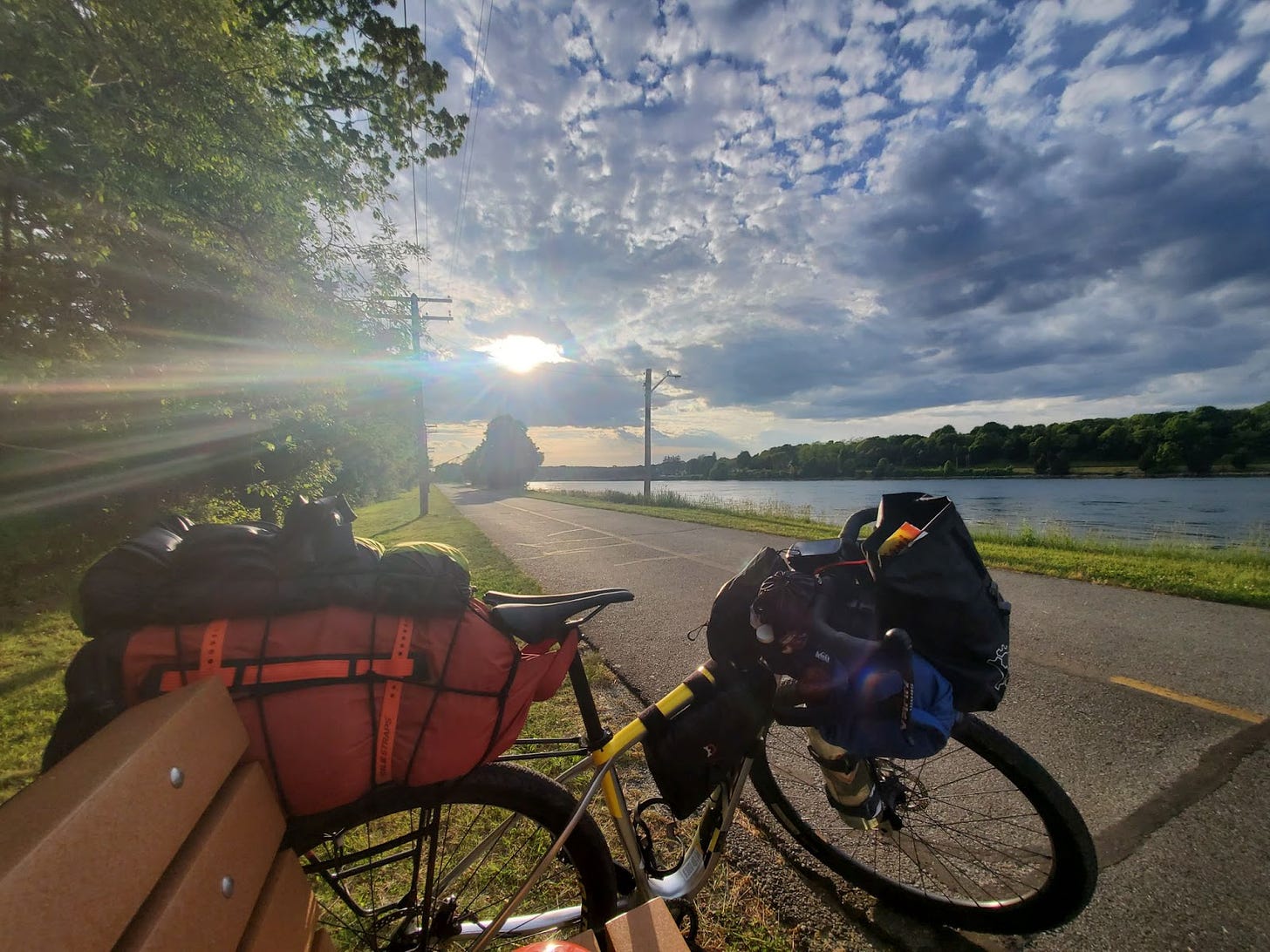 A bicycle parked in front of a bench next to the road. A lake is across the road, and the sun is shining low in the sky through clouds.