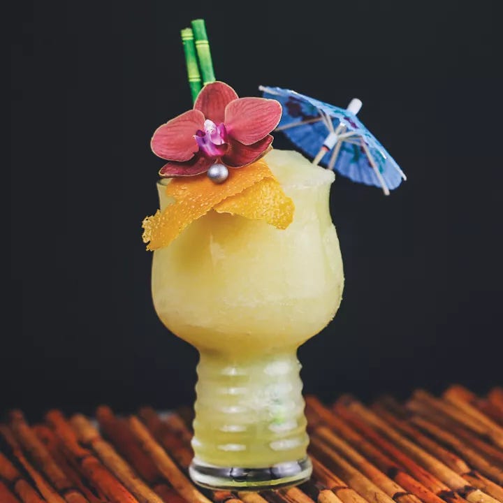 A glass Tiki mug sits on a bamboo mat. The glass is filled with a yellow slushy drink, and is garnished with an orchid, a paper umbrella, and an orange peel.