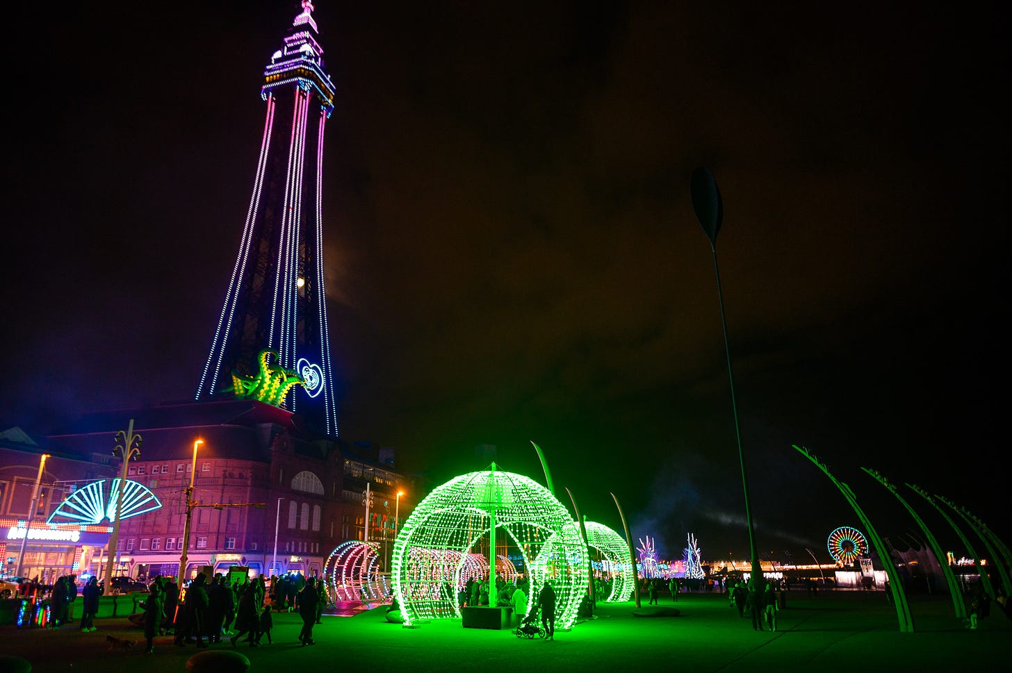 People walk through domes of green lights on the promenade in Blackpool. The tower is lit up.