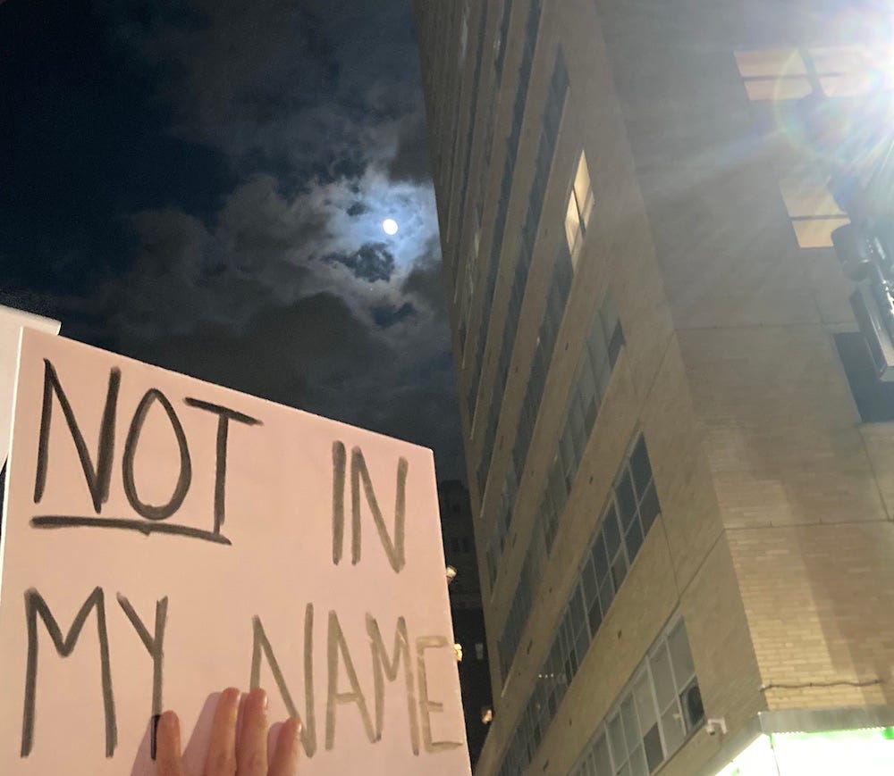 A photo taken looking up into the night sky in a city. Behind a thin cloud, the full moon shines. Below, someone holds a sign reading “NOT in my name.”