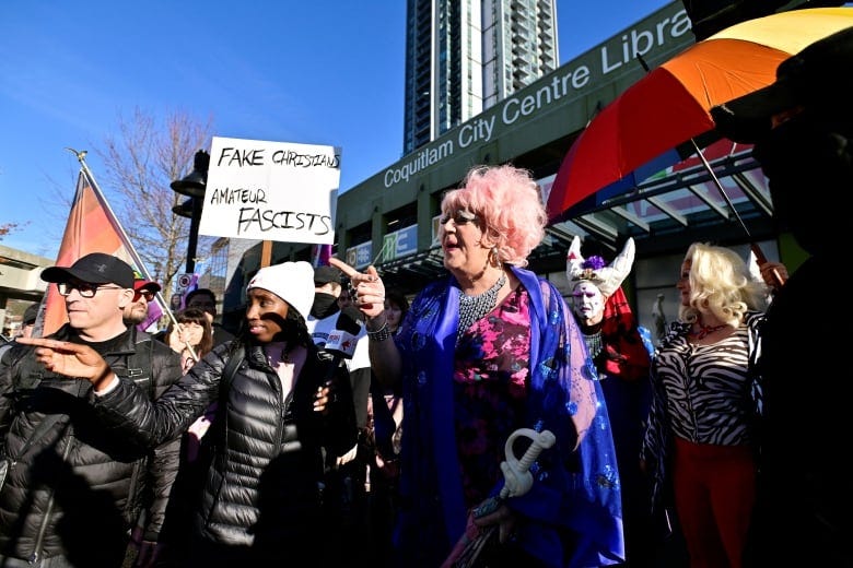 A drag queen, surrounded by supporters, points to the left of the frame outside a public library. A sign in the background reads 'Fake Christians Amateur Fascists'.