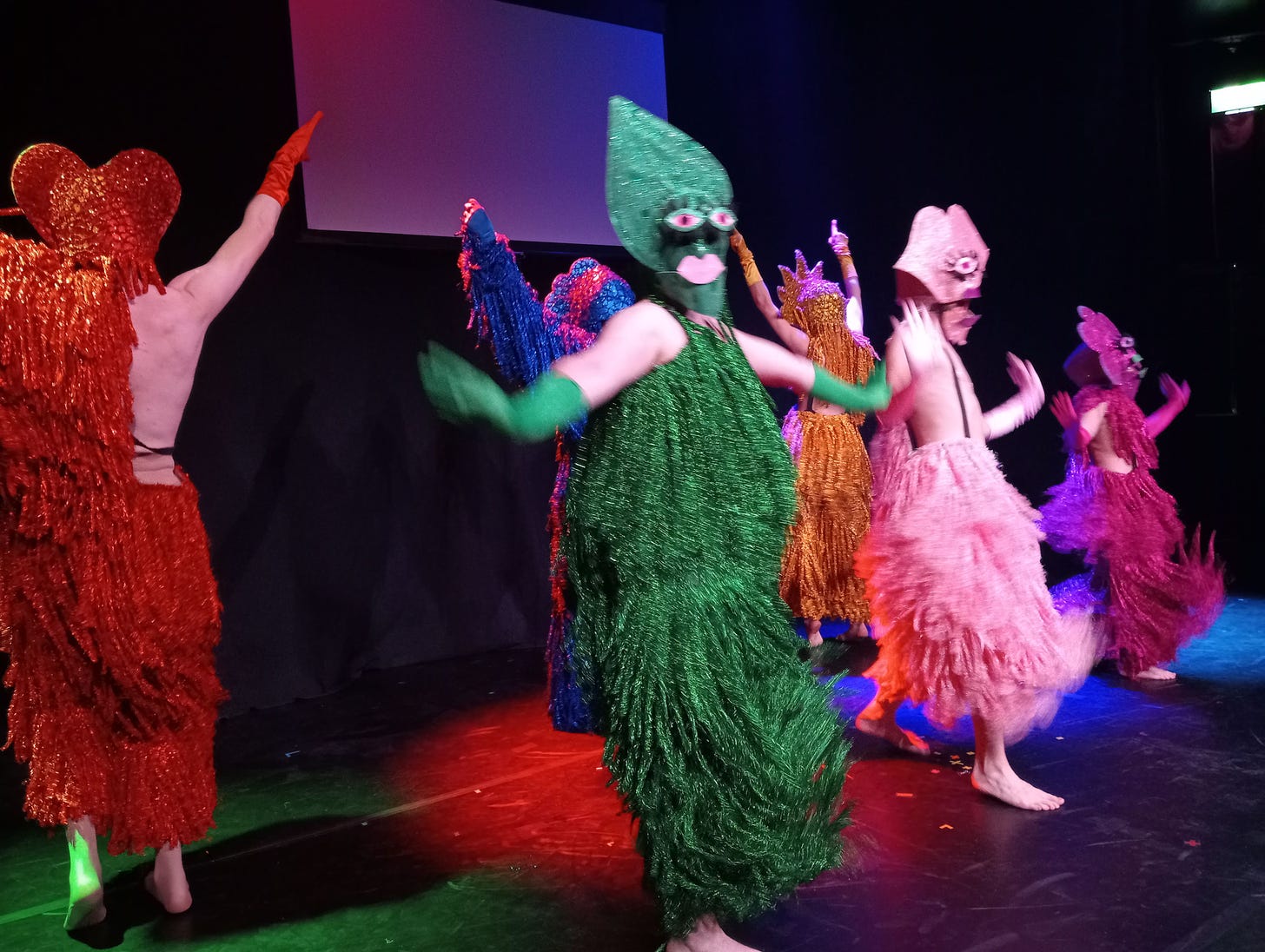 An image of 6 drag performers in matching costumes, each a different colour (red, green, pink, blue, orange and purple). Sparkly and cartoon-like.