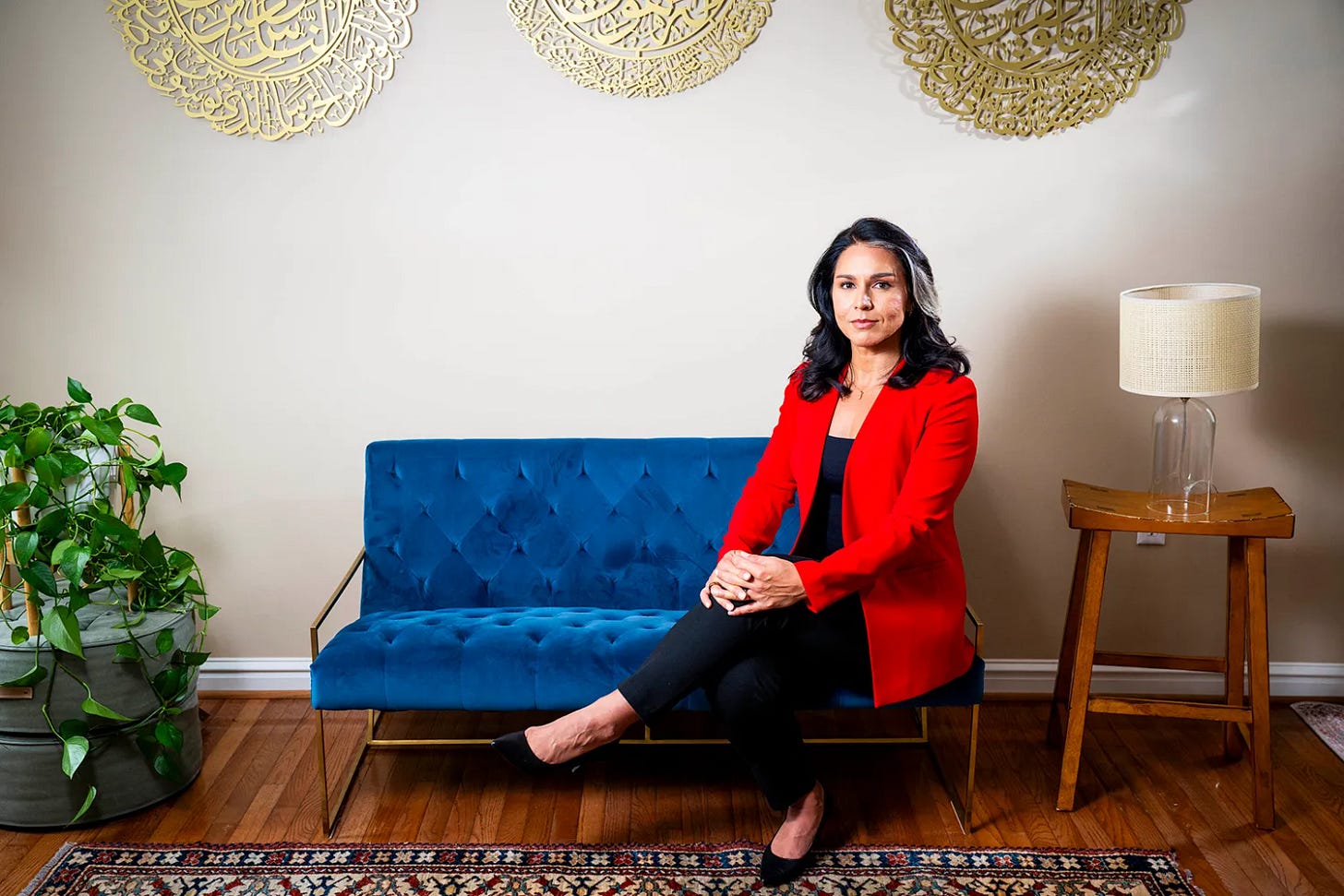 Tulsi Gabbard in a red suit jacket sits on a small blue couch with her hands clasped on her knee. Behind her are golden circular wall hangings with words in a foreign text on them. A small table with a lamp is on one side, on another an ottoman with a plant atop it.