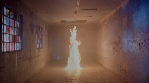 In a long interior space with concrete walls and colored glass blocks along the left wall, a figure in the middle is engulfed in flames towards the end of the hall.