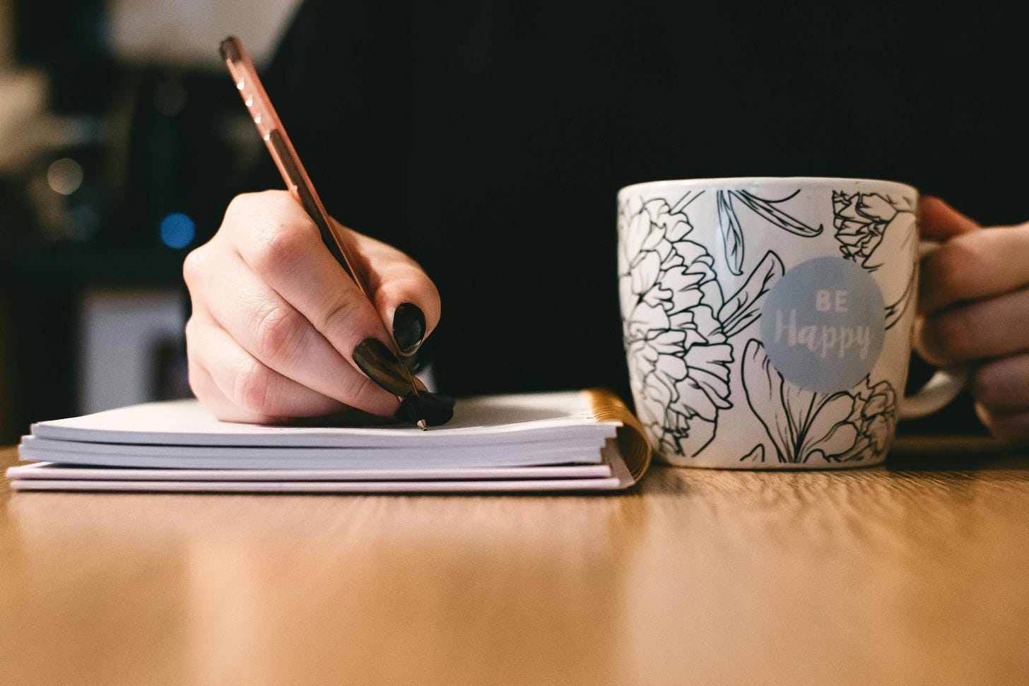 person wearing black writing in notebook with mug that says "be happy" in other hand