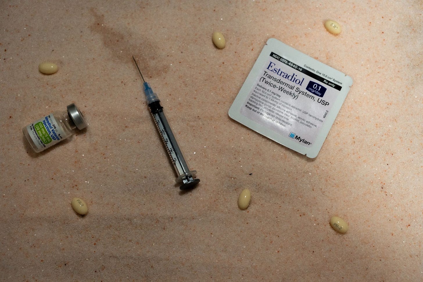 On a bed of pink salt lays estrogen vials, patches, needles, and progesterone capsules. The estrogen vial and needle are sadly empty.