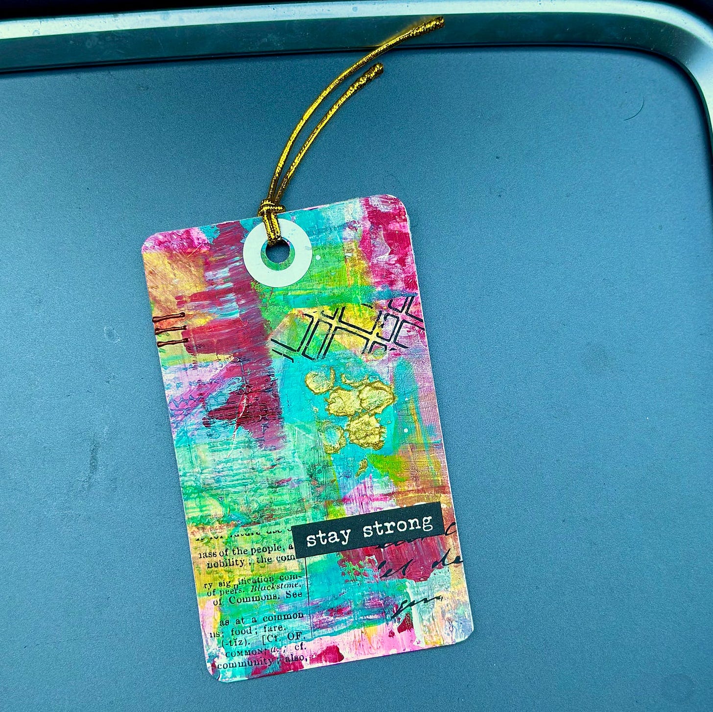 Mixed media tag with colorful bursts of acrylic paint punctuated with textured gold stenciling and the words “stay strong”
