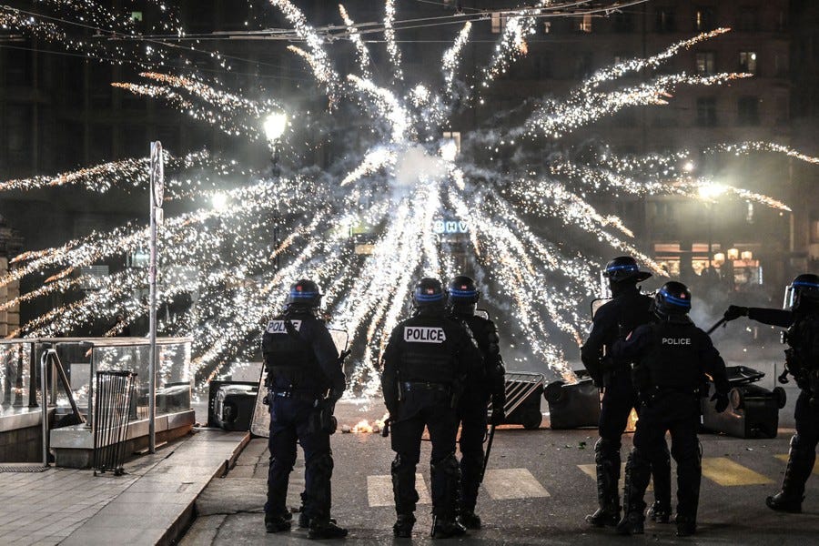Police officers stand in a street in front of an exploding firework.