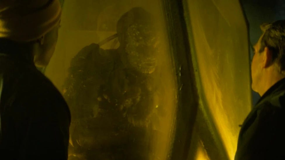 This image shows Ryan Sinclair (played by Tosin Cole, left) and Graham O'Brien (played by Bradley Walsh, right) talking to Stenza warror T'Zim-Sha (played by Samuel Oatley, center) as he begins freezing inside the stasis chamber in which Graham has locked him. Ryan's and Graham's backs are to camera in this still photo from "The Battle of Ranskoor Av Kolos" (Doctor Who Series 11's tenth episode and season finale). This image shows Ryan Sinclair (played by Tosin Cole, left) and Graham O'Brien (played by Bradley Walsh, right) talking to Stenza warror T'Zim-Sha (played by Samuel Oatley, center) as he begins freezing inside the stasis chamber in which Graham has locked him. Ryan's and Graham's backs are to camera in this still photo from "The Battle of Ranskoor Av Kolos" (Doctor Who Series 11's tenth episode and season finale).