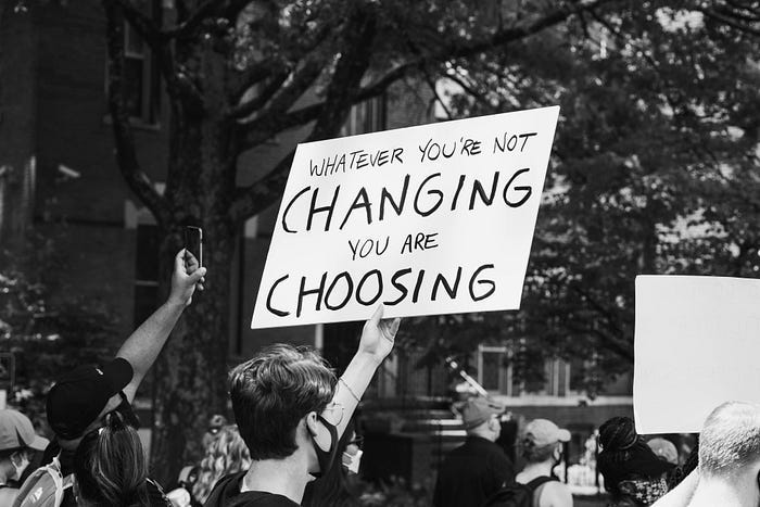 A masked protestor holds up a sign that reads, “Whatever you’re not changing, you are choosing.” A masked protestor holds up a sign that reads, “Whatever you’re not changing, you are choosing.”