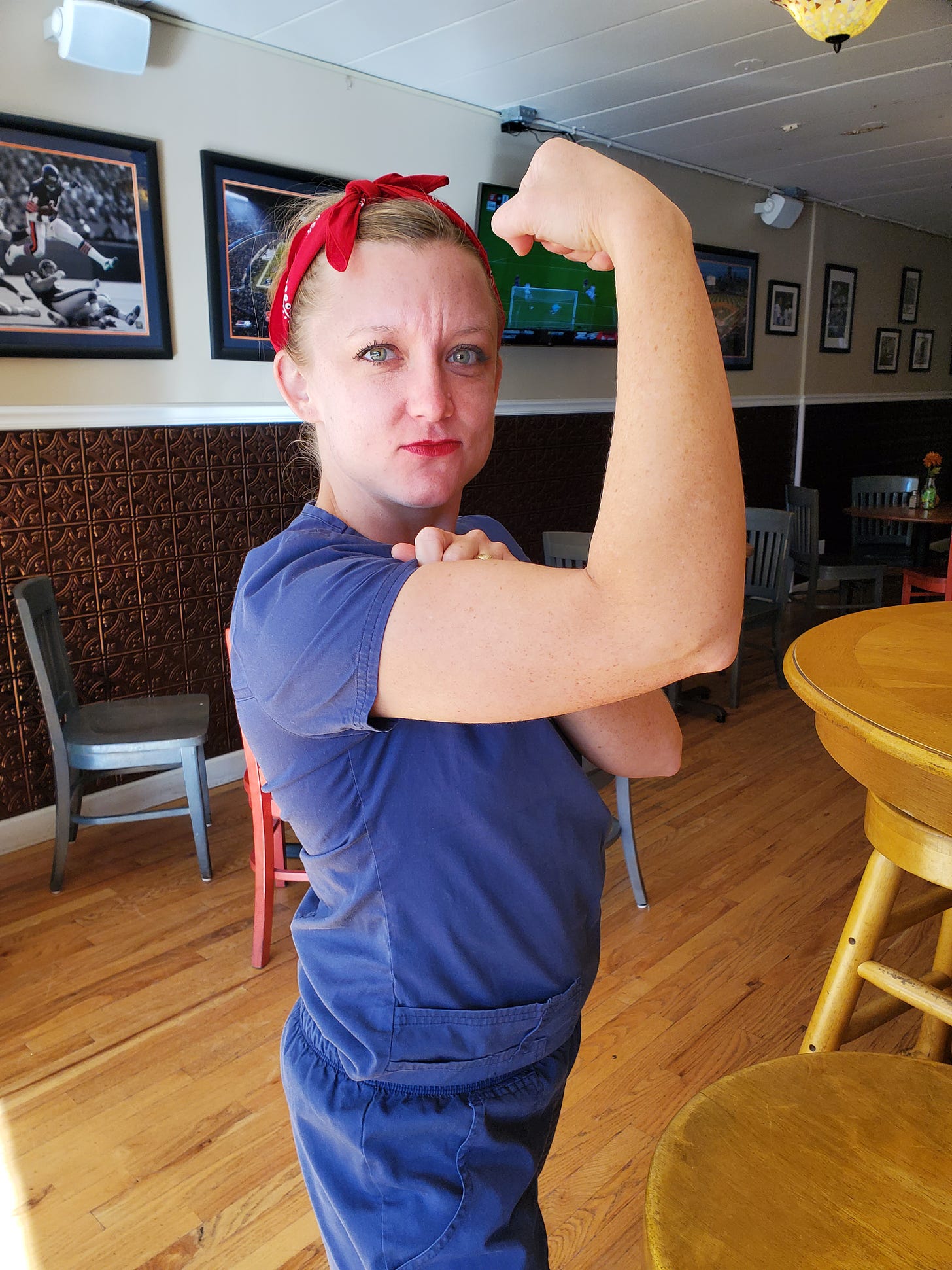 Woman wearing red bandana and blue scrubs flexing right bicep staring powerfully at the camera