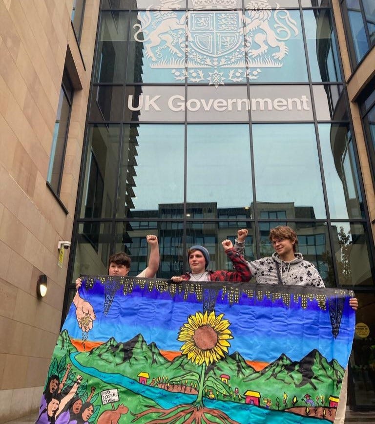 Three youth activists holding a banner with a picture of a flower against the blue. They are standing in front of the UK government building