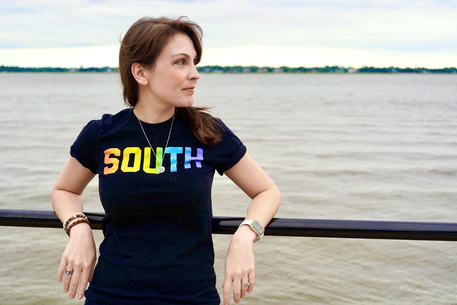 A photo of Kendra, a white woman with brunette hair, with her back against a railing with water behind her. She’s wearing a black t-shirt with the word South on the front. She’s turned her head and is looking off to the right.