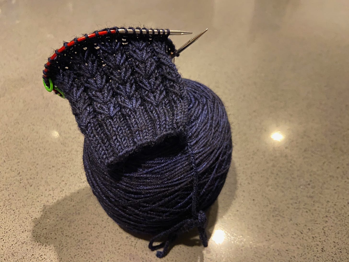 One Constellate Mitt, which is partially knitted and placed on top of a skein of yarn and is placed on a kitchen counter. The yarn is a dark blue with black coloring.