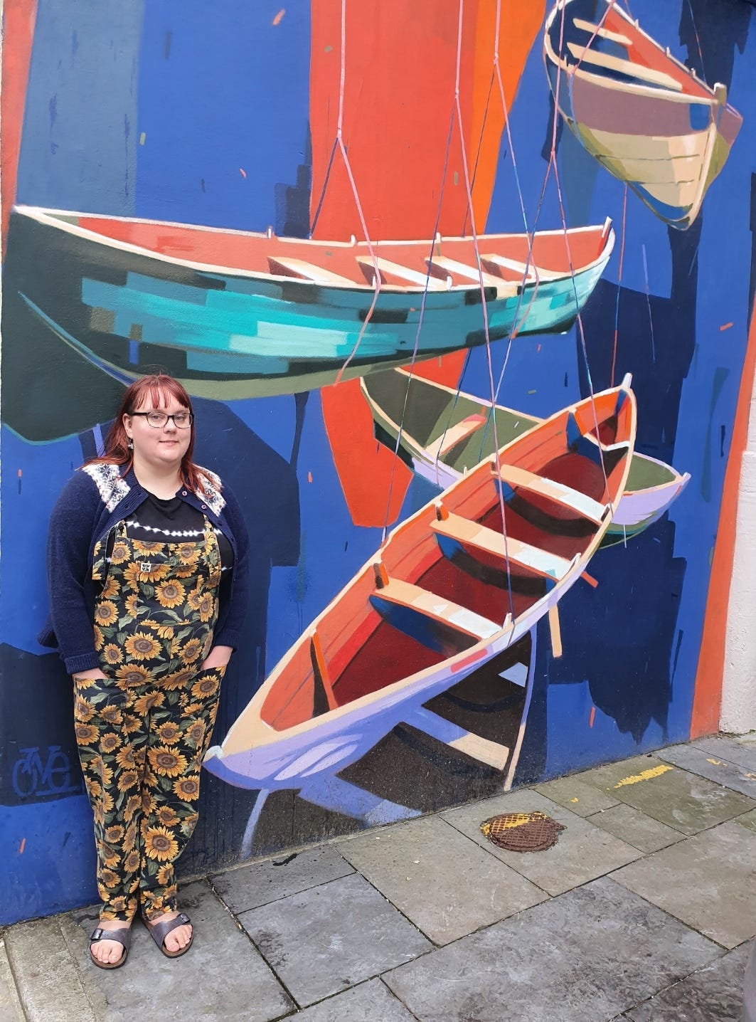 Hannah, aged 34, has dyed red hair, a blue cardigan with a Fair Isle yoke pattern around the neck, a black and white tie dye effect t-shirt, sunflower patterned dungarees, glasses with thick blue frames, and iridescent Birkenstock sandals. She is standing in front of a mural with a blue and orange background and three rowing boats in various colours in its foreground. 