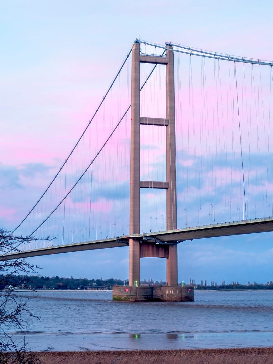 Photograph of one of the tall support towers of the Humber Bridge. There is a blue and pink cloudy sky just before sunset.