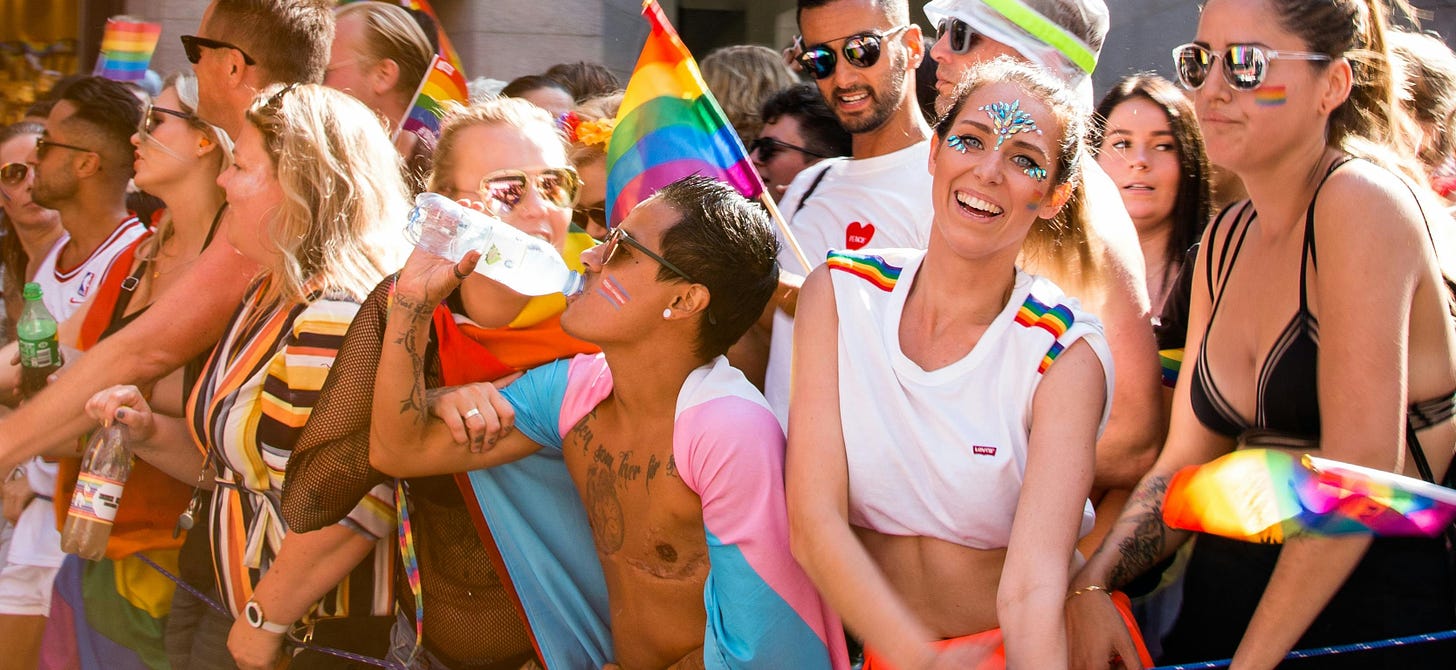 A raucous, crowded Pride celebration with many rainbow flags. Centered is a trans man, top surgery scars proudly showing, draped in the trans flag.