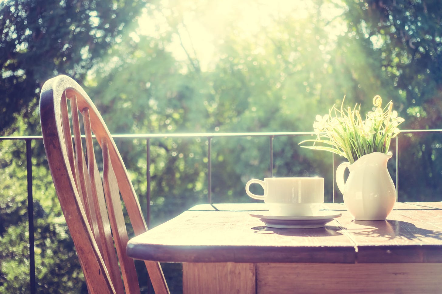 Close up of a chair beside a table in the morning. On the table there's a cup of coffee and a white jar filled with white flowers and green leaves.