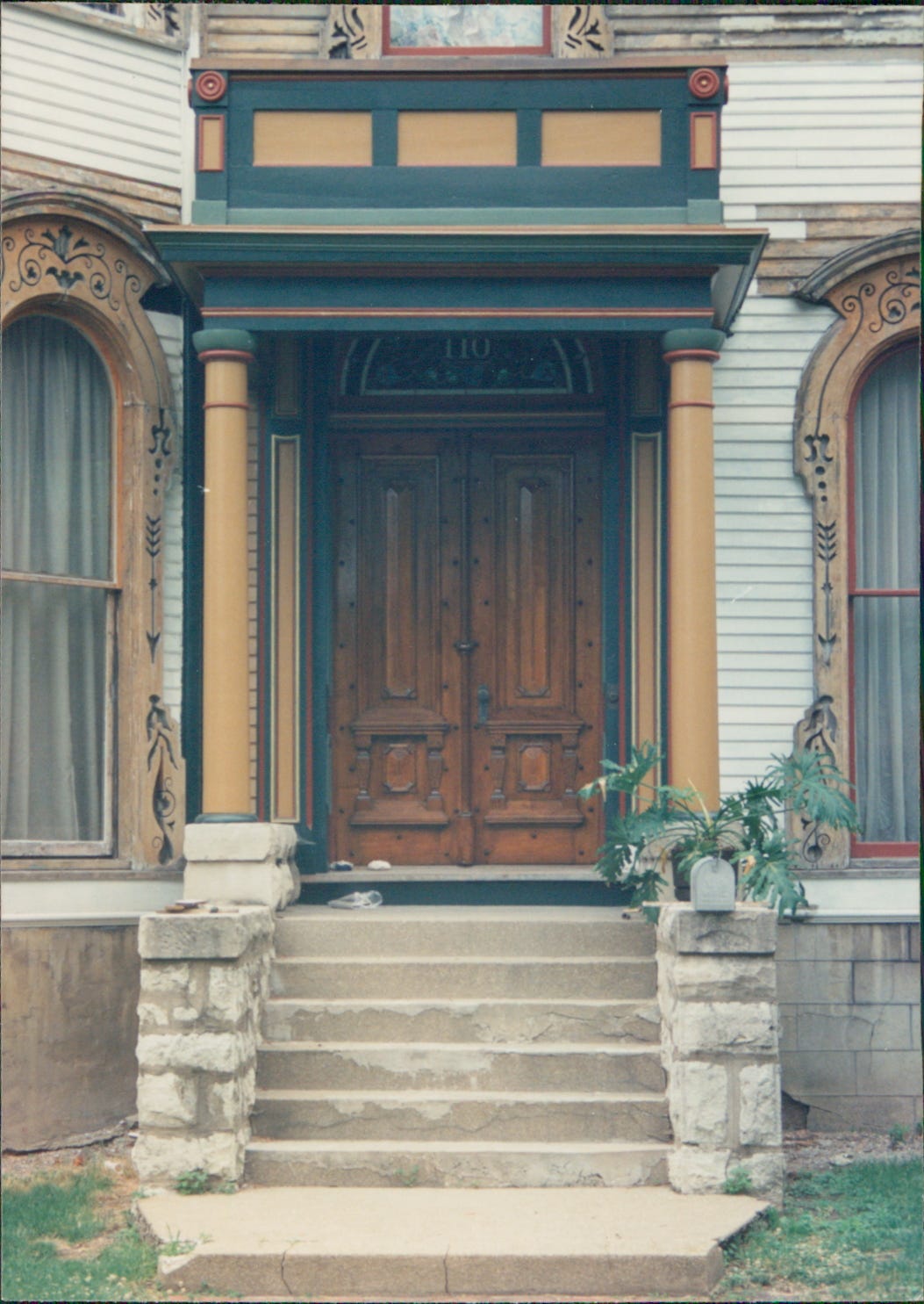 Front entrance of Second Empire Victorian home with ornate walnut double entryway doors framed by columns and roof. The woodwork around the entrance is painted Victorian yellow, colonial red, shutter green, and olive.