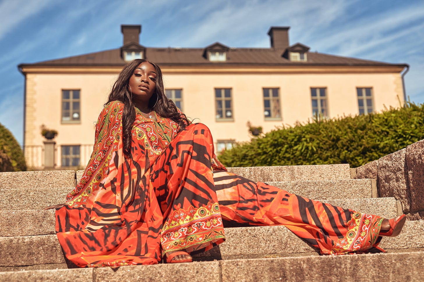 Lovette Jallow vibrant orange traditional clothing sits smiling on stone steps in front of a European building, embodying grounded joy and self-containment. Lovette Jallow vibrant orange traditional clothing sits smiling on stone steps in front of a European building, embodying grounded joy and self-containment.