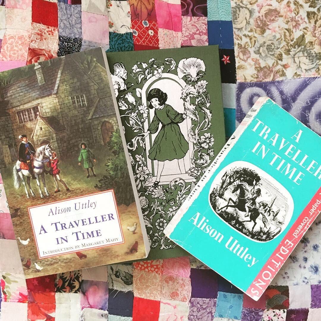 Three copies - one hardback in green, and two paperbacks - of Alison Uttley’s a Traveller in Time. The books are resting on a piece of patchwork in pink, purple and blue. 