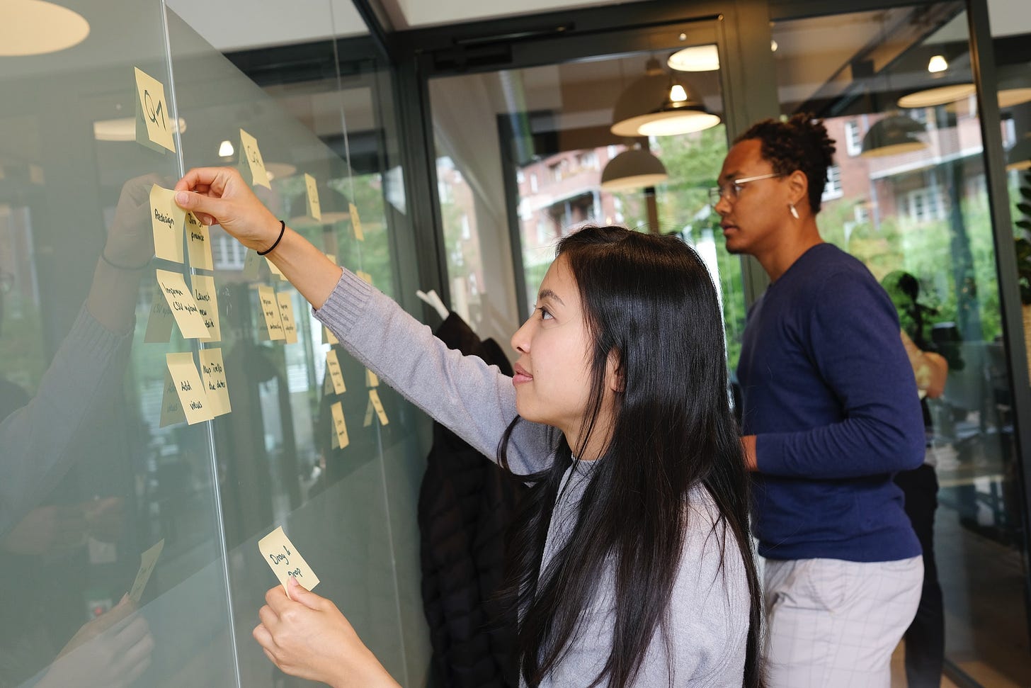 image of woman in light purple shirt in foreground placing sticky notes on glass wall. a man wering glasses is in the background looking at the same wall.