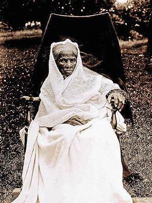 Harriet Tubman sits in a chair outside with a white blanket draped over her legs that extends to the ground. A white shawl is draped over upper body and head her face and some of her gray hair are visable. 