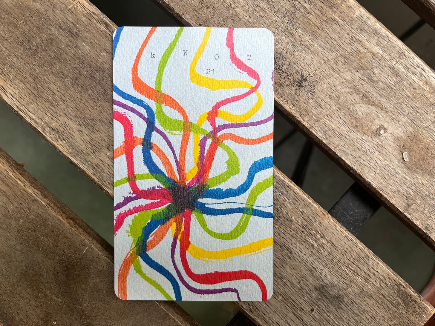 The card "Knot" lies on a wooden picnic table. There are gaps between each wooden slat, so you can see the ground in the background. The card is a painting of many lines of different colors coming together at the center. The word "knot" as well as the number 21 are printed on the card and seem to have been written with a typewriter. 