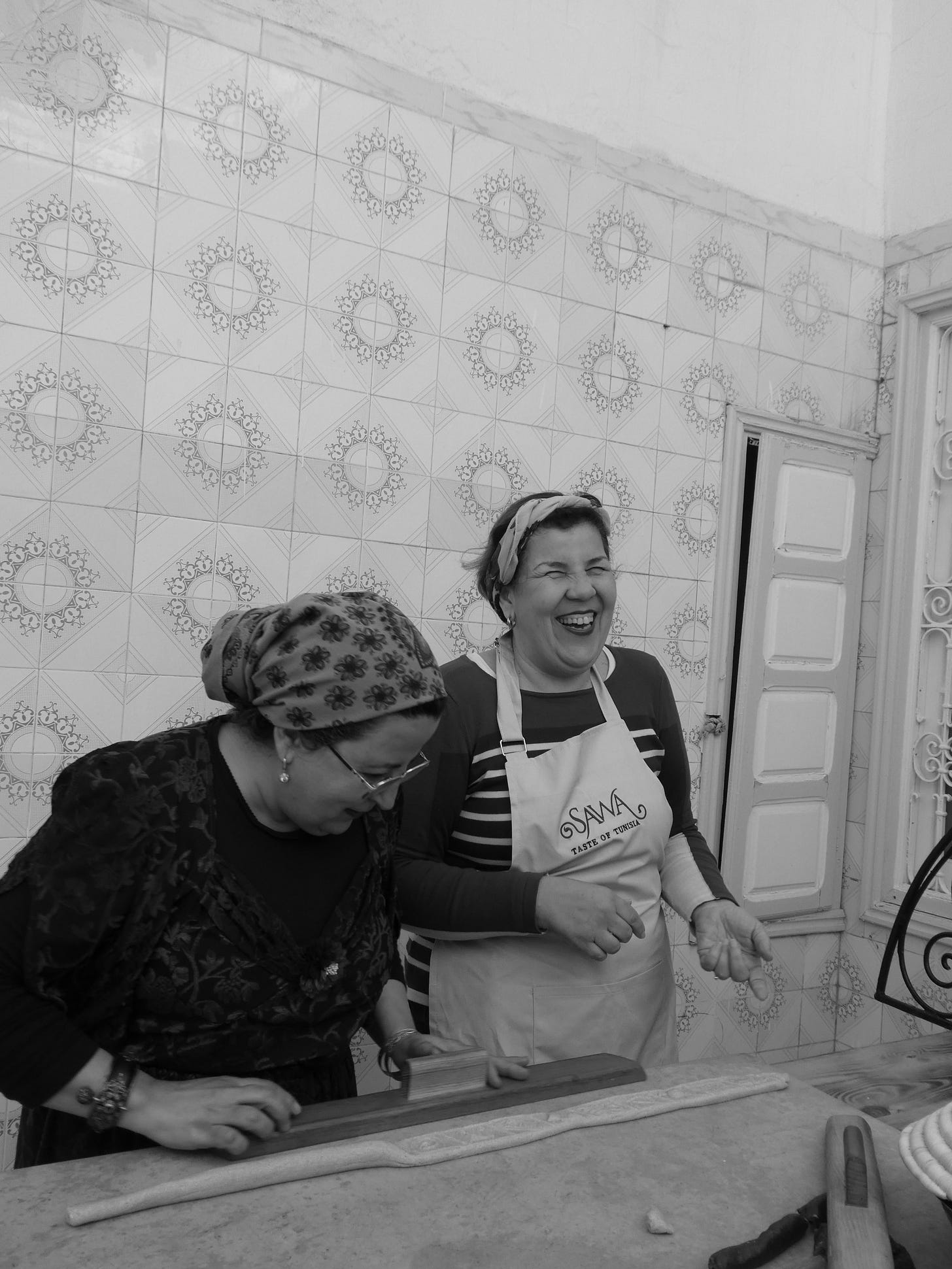 In front of a tiled wall halima uses a mould whilst lamia laughs