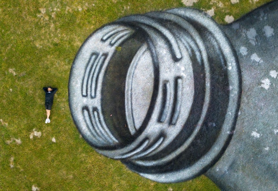 An aerial view of a person lying on their back on grass, beside a huge painted mural of a plastic bottle.