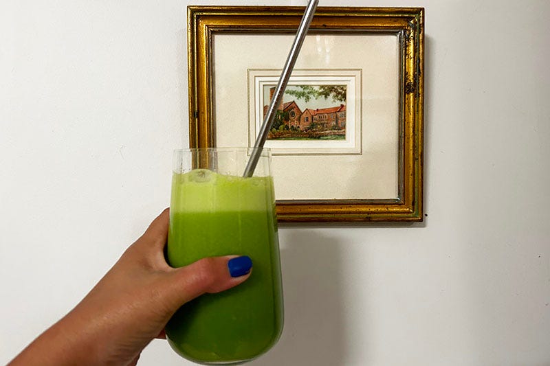 Hand holding a glass of green juice against a small oil painting.