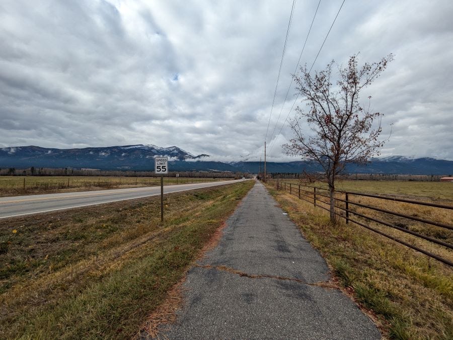 a long empty stretch of road with mountains at one end and empty fields on both sides. A walking path is along one side.