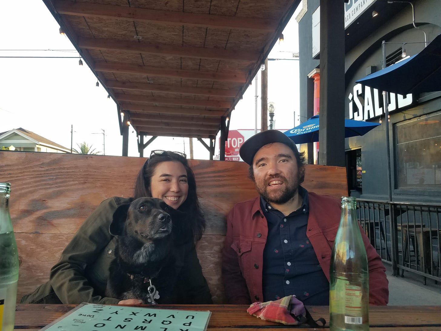 A smiling man with brown hair, short beard, brown baseball cap, and red jacket seated next to a smiling women with black hair and a green jacket, at an outdoor restaurant booth. The woman has a cute black lab mix dog in her lap. There is a letterboard on the table.