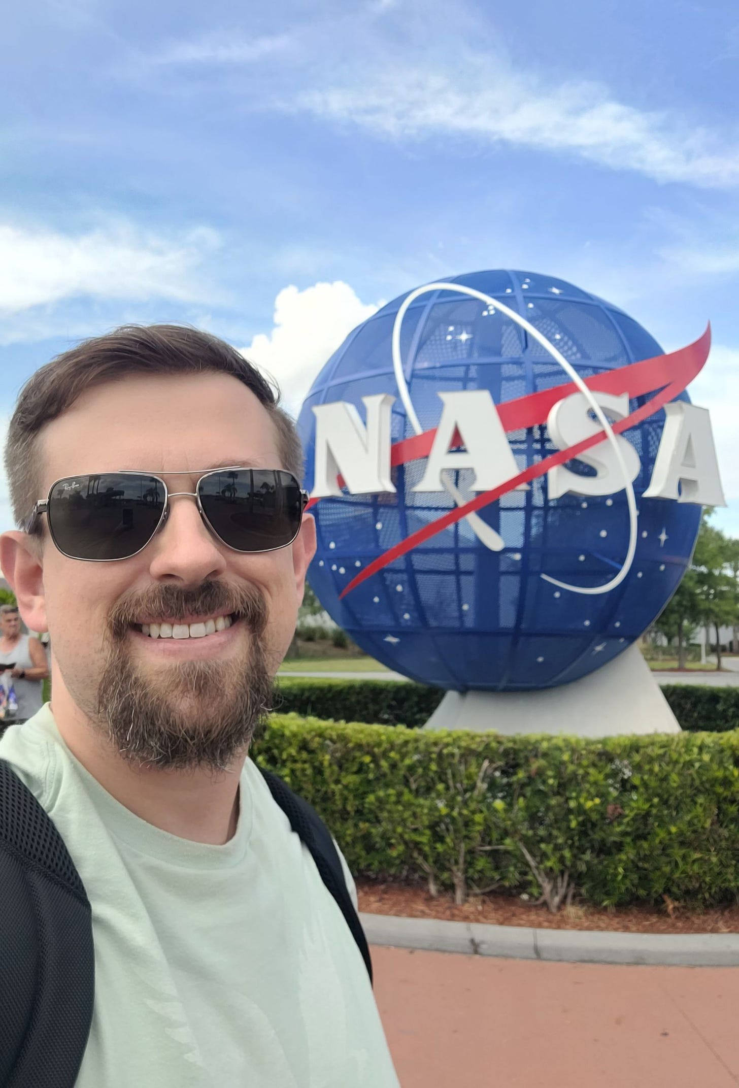 Eu, homem branco e loiro de cavanhaque, com óculos escuros, camiseta verde e alças de mochila, estou na frente do símbolo da NASA, um globo azul com o nome NASA em branco na frente e traços brancos e vermelhos. O dia está claro e dá pra ver o céu.