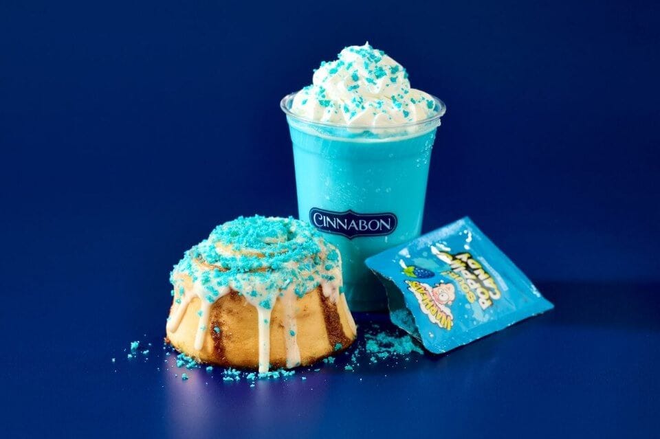 A cinnamon roll with blue sour candy sprinkled on top next to a neon blue drink with sour candy sprinkled on top and a torn open Warheads package