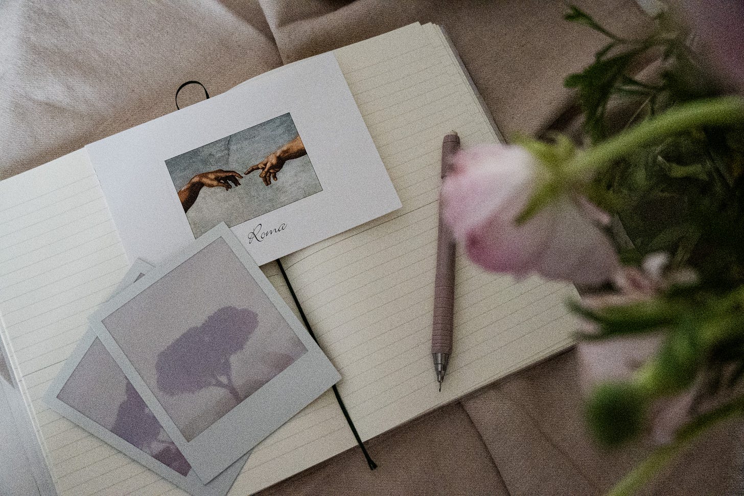 Softly lit writing desk with an open notebook, vintage-style Rome postcard and polaroids, a blush pink pen, and a blurred rose in the foreground. Softly lit writing desk with an open notebook, vintage-style Rome postcard and polaroids, a blush pink pen, and a blurred rose in the foreground.