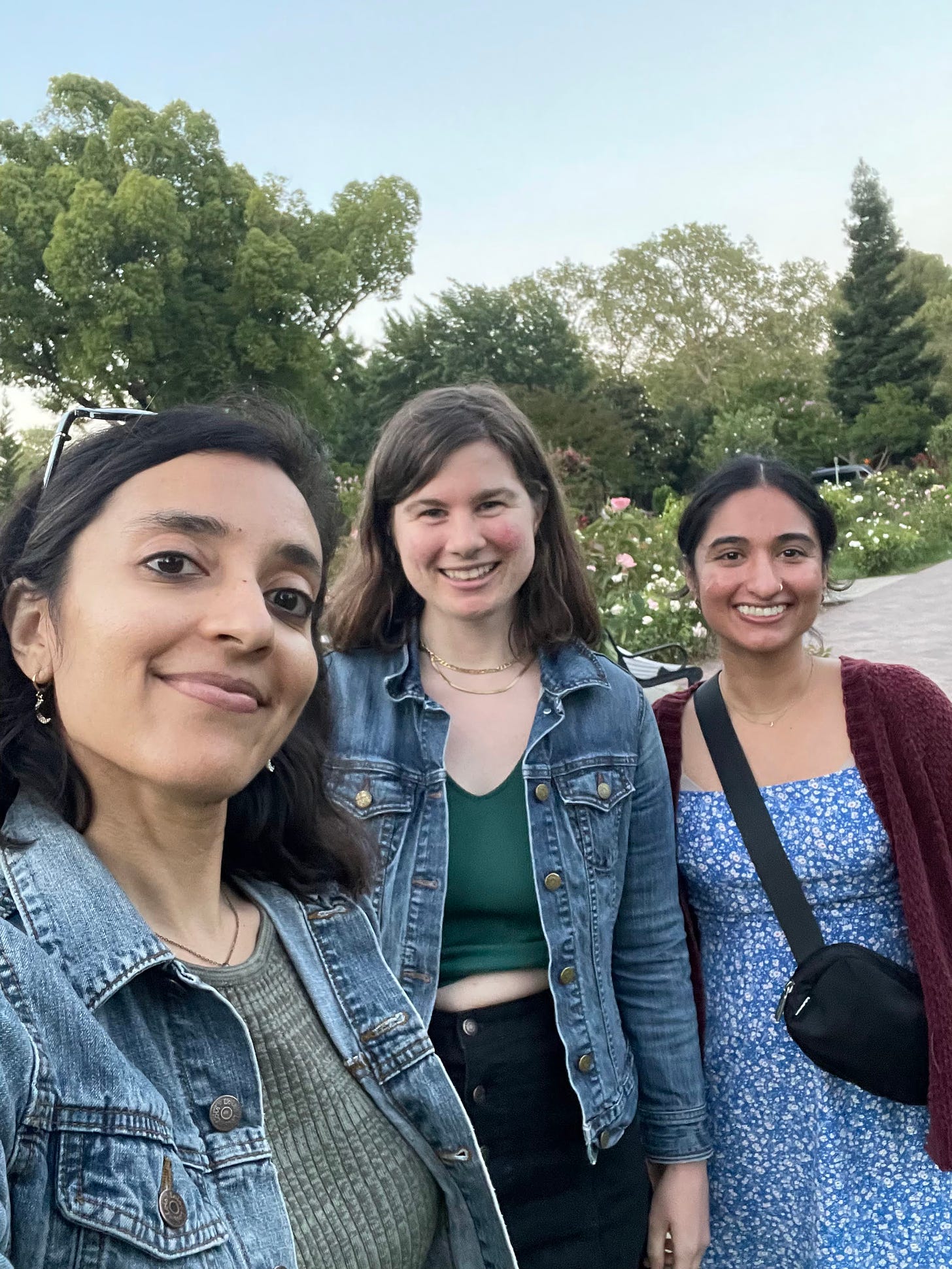 Three women, stand in a rose garden and smile into the camera. The woman on the right has her hair in a ponytail and wears a red cardigan atop a blue dress. The other two women wear jean jackets over green tops.