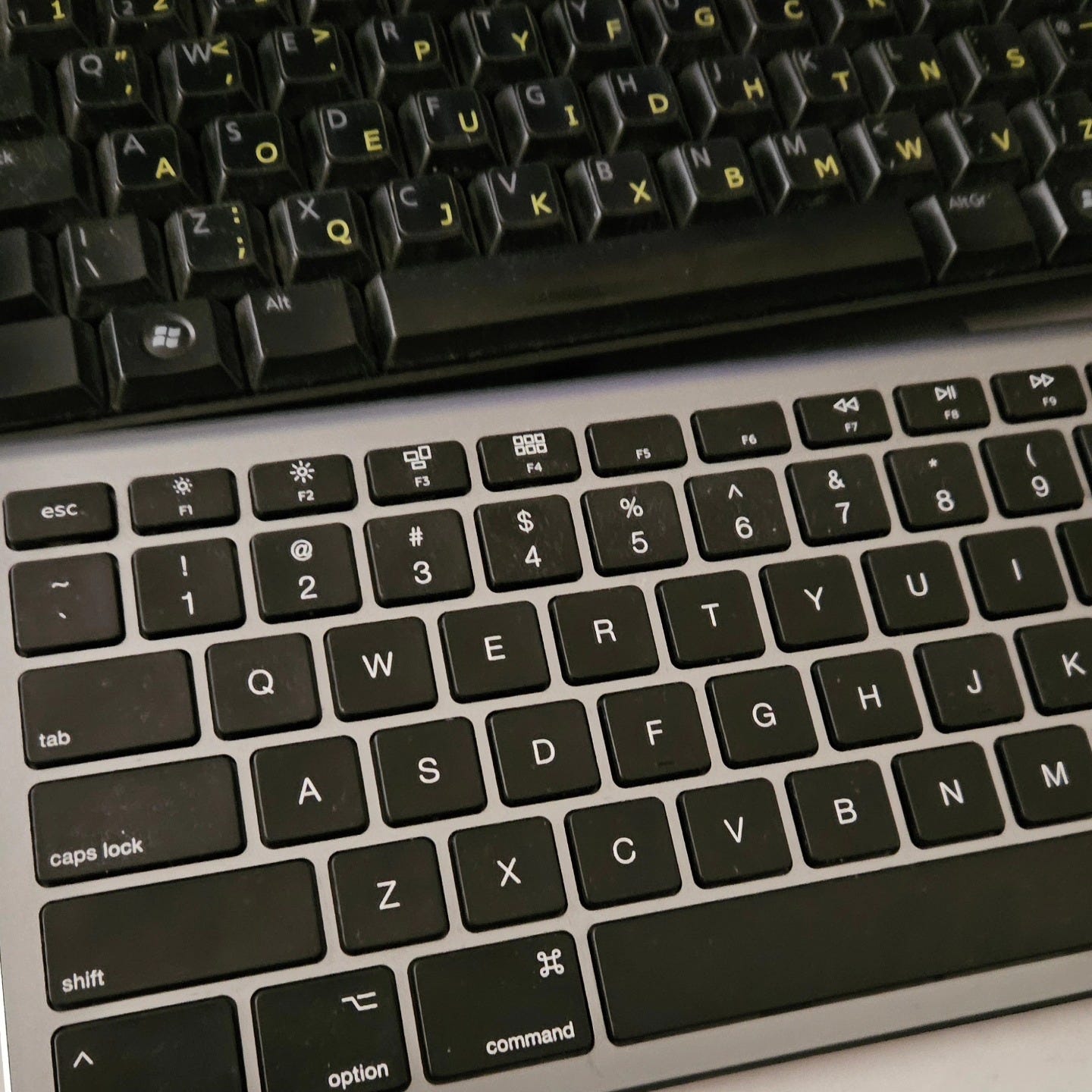 Image of two keyboards on Damon Lord's desk, the top is a black Windows keyboard with Dvorak layout on it in yellow, and the lower keyboard is a silver and black standard Mac keyboard.