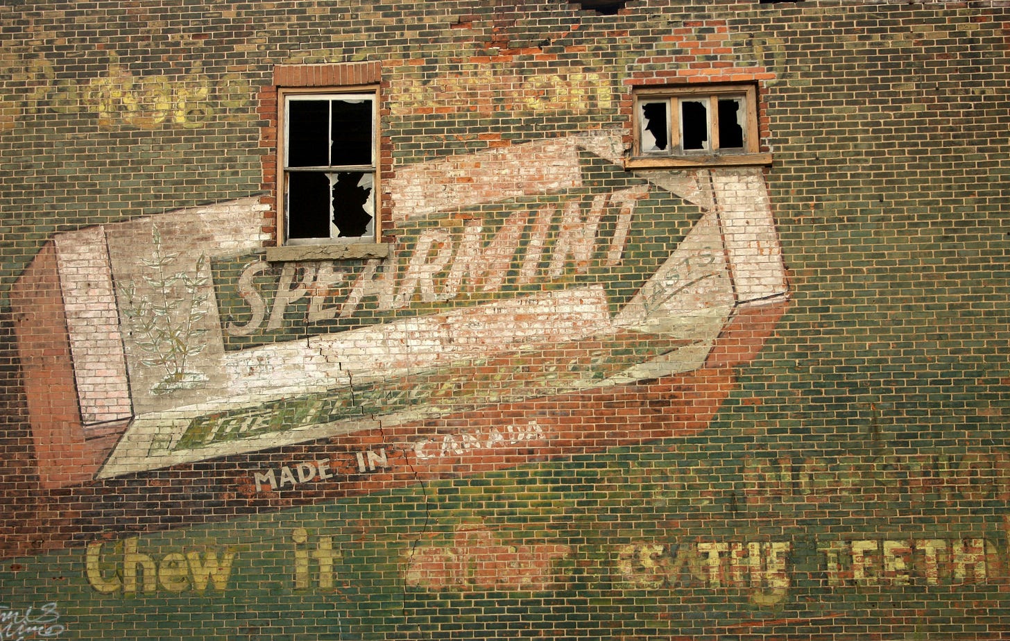 An old, faded ad for Wrigley's spearmint gum painted on a brick wall pierced by two broken windows.